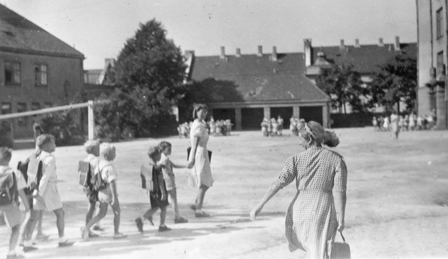 Foto: Første skoledag i august 1947 Første skoledag i 1947. Elevene taues til klasserommet av frøken. Det er seks gutter som går bak en ung dame i kjole i skolegården.