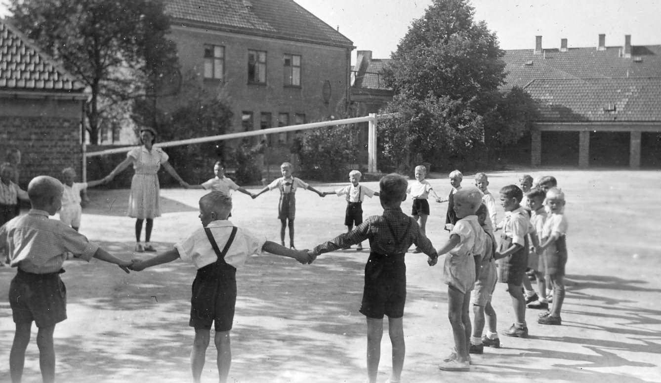Lek i skolegården på Rosenhof skole. Gutter i kortbukser som holder hender med en lærerinne i kjole. Bildet er fra 50-tallet.