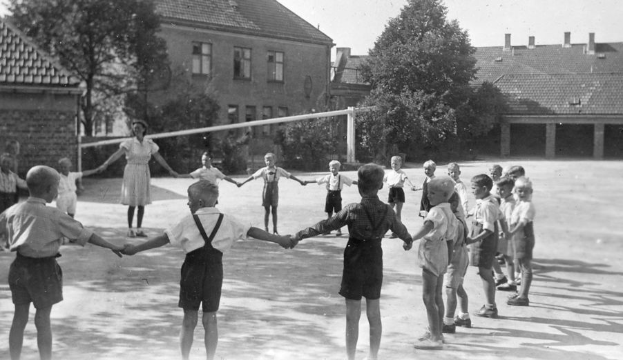 Foto: Lek i skolegården Lek i skolegården på Rosenhof skole. Gutter i kortbukser som holder hender med en lærerinne i kjole. Bildet er fra 50-tallet.