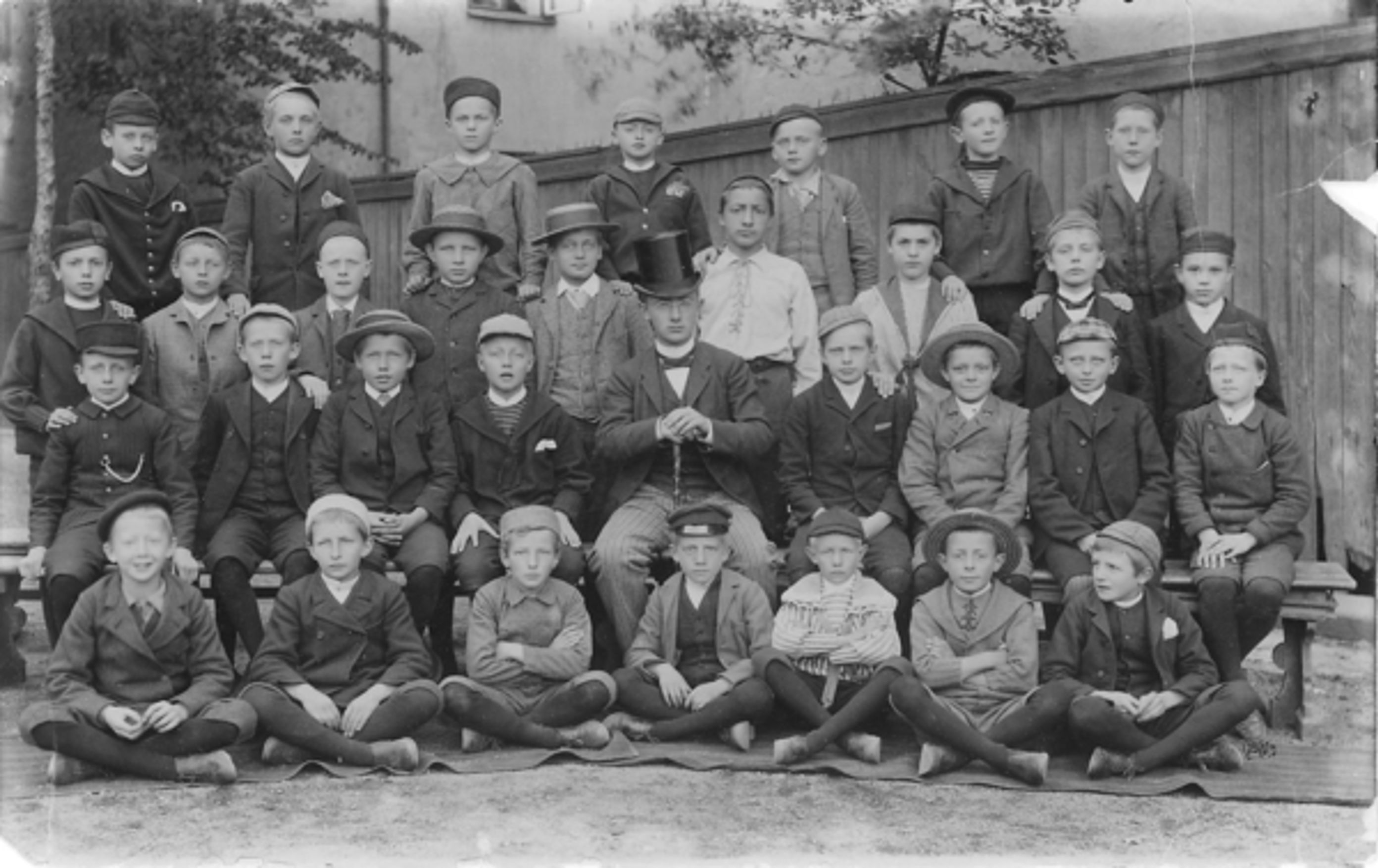 Bilde av en gutteklasse på Borgerskolen i 1890. Alle har på seg lue eller hatt. Læreren har på flosshatt.