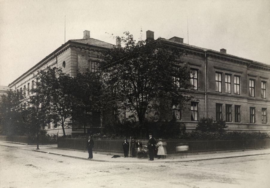 Foto: Christiania Borger- og Realskole i 1905. Fotograf: Jespersen, Jesper. Oslo Museum/OB.F18703 Bilde av Borgerskolen i 1905. Det er to menn i dress og en dame i kjolet på bildet samt to barn.
